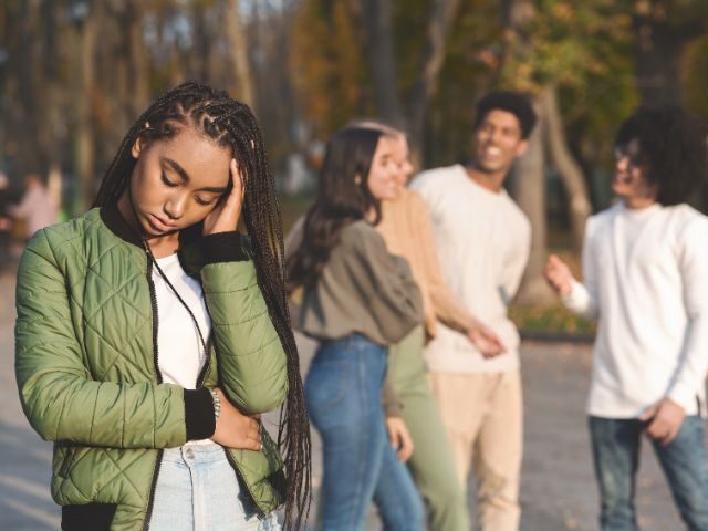 Black female teen holding her stomach and touching her head, avoiding interaction with a group of other students